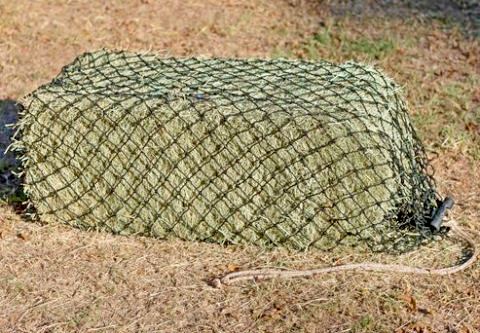 Small bale cinch hay net for horses filled with a small two-string bale of grass hay and laying on the ground.